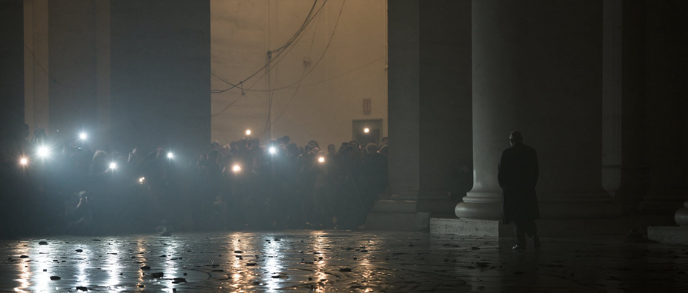 A crowd of press lights under the colonnade at night, one figure walking away