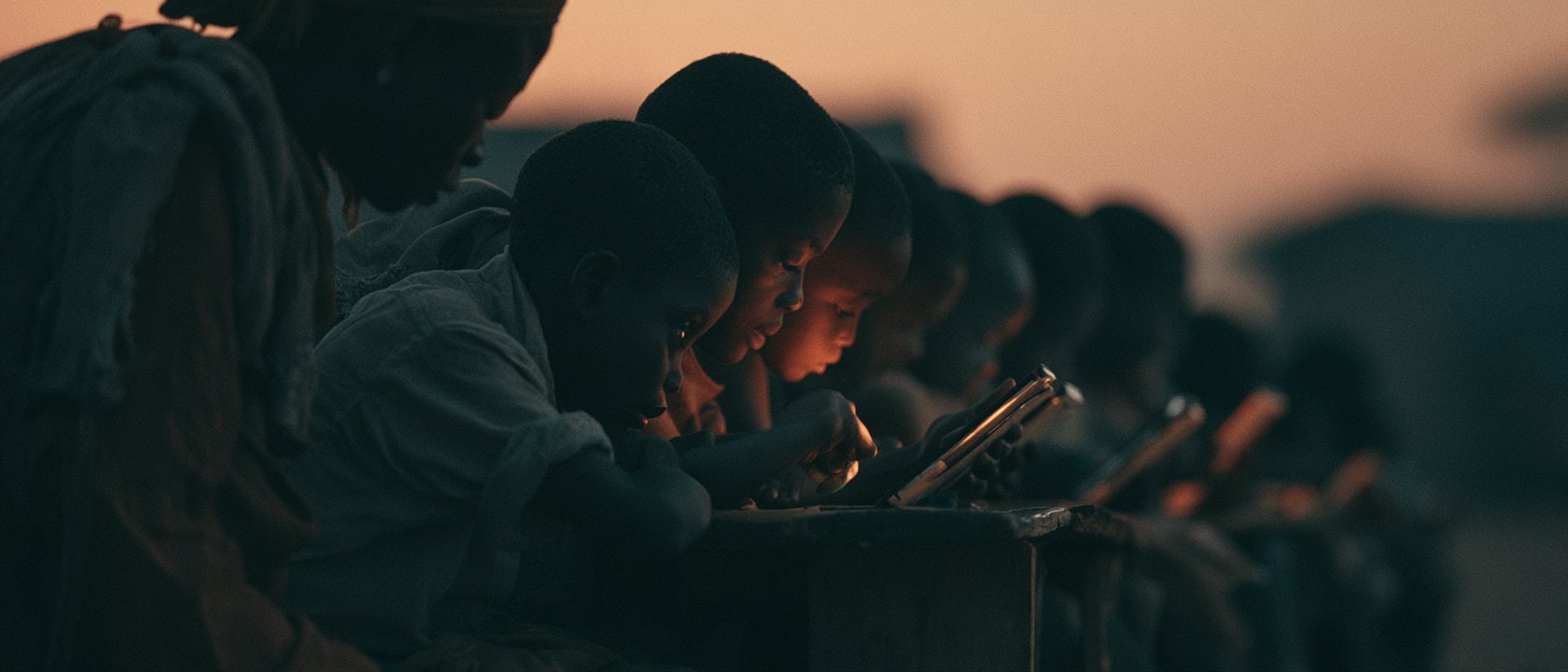 Children at rough desks at sunset, faces lit by tablet screens, a teacher leaning close